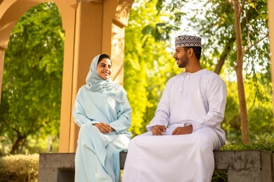 مفهوم العلاقات الزوجية الناجحة السعيده، قضاء عطلة نهاية الاسبوع في أحد الحدائق ، التنزه في الهواء الطلق ،زوجان عربيان خليجيان عمانيان يرتديان الزي التقليدي يقومان بتبادل الأحاديث والدردشات، اجواء مليئة بالمحبة والود