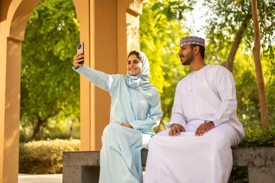التقاط صور سيلفي تذكارية عبر الهاتف المحمول، التنزه في الهواء الطلق، مفهوم العلاقات الزوجية الناجحة السعيده، قضاء عطلة نهاية الاسبوع في أحد الحدائق ،زوجان عربيان خليجيان عمانيان يرتديان الزي التقليدي يقومان بتوثيق اللحظات الجميلة