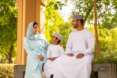 حديقة ومنتزه عائلي ترفيهي، قضاء الوقت الممتع مع الأسرة, الاستمتاع بالنزهة في الهواء الطلق, تقوية وتعزيز الروابط الأسرية، تبادل الأحاديث بإيماءات السرور، عائلة عربية خليجية عمانية يرتدون الزي التقليدي يجلسون في إحدى الحدائق العامة في النهار