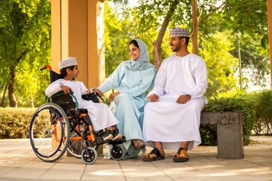 The concept of a happy family, spending enjoyable family time, an Omani boy wearing a dishdasha and kumma sitting in a wheelchair in one of the parks with his parents, gives positive energy to patients with mobility disabilities.
