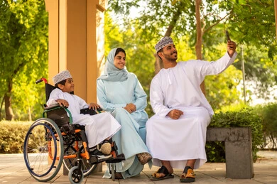 Spending enjoyable family time, an Omani boy wearing a dishdasha and kumma sitting in a wheelchair in one of the parks with his parents, giving positive energy to patients with mobility disabilities, a father taking a group family selfie with a mobile phone.