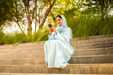 Integrating technology into daily life, gestures of happiness and joy, an Omani woman browsing social media, a Gulf Arab Saudi woman wearing an abaya and hijab sitting outdoors holding her mobile phone.