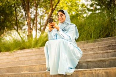 استخدام جهاز تقني حديث ، ايماءات السعادة و السرور، سيده عمانية تقوم بتصفح مواقع التواصل الاجتماعي، امراة عربيه خليجيه سعودية ترتدي العباءة و الحجاب تجلس فى الهواء الطلق تمسك بيدها الجوال