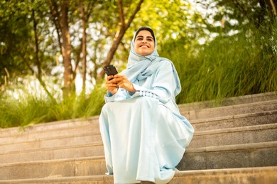 النظر الى شيء ما، ايماءات السعادة و السرور، سيده عمانية تقوم بتصفح مواقع التواصل الاجتماعي، امراة عربيه خليجيه سعودية ترتدي العباءة و الحجاب تجلس فى الهواء الطلق تمسك بيدها الجوال