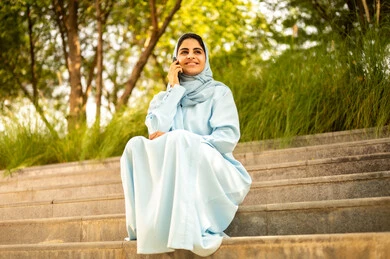 ايماءات السعادة و السرور، سيده عمانية تقوم بإجراء مكالمة هاتفية باستخدام الهاتف المحمول، استخدام الأجهزة والتقنيات الحديثة، امراة عربيه خليجيه سعودية ترتدي العباءة و الحجاب تجلس فى الهواء الطلق تمسك بيدها الجوال