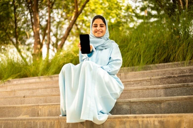 The use of modern devices and technologies, an Omani woman displaying a blank black screen, a Saudi Arabian Gulf Arab woman wearing an abaya and hijab sitting outdoors holding a mobile phone, gestures of happiness and joy.