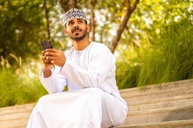 تصفح مواقع التواصل الاجتماعي ،تبادل الأحاديث مع الأهل والأصدقاء، رجل عربي خليجي عماني يرتدي الدشداشة و الكمة يجلس فى الهواء الطلق يمسك بيدة الجوال و يقوم بالنظر الى شيء ما