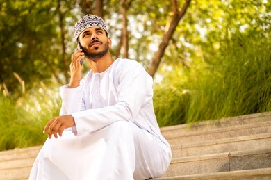 تبادل الأحاديث مع الأهل والأصدقاء، إجراء مكالمة هاتفية باستخدام الهاتف المحمول، رجل عربي خليجي عماني يرتدي الدشداشة و الكمة يجلس فى الهواء الطلق يمسك بيدة الجوال و يقوم بالنظر الى شيء ما