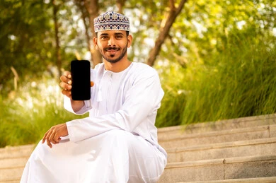 استخدام الأجهزة والتقنيات الحديثة، عرض شاشة سوداء فارغة، المنتزهات والحدائق العامة، رجل عربي خليجي عماني يرتدي الدشداشة و الكمة يجلس فى الهواء الطلق يمسك بيدة الهاتف المحمول، ايماءات السعادة و السرور