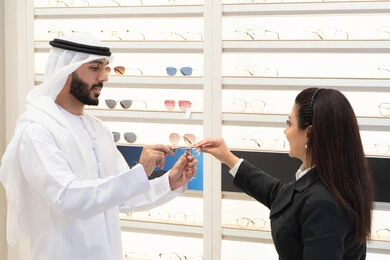 Emirati woman helping customers choose the right size, admiring eyeglasses while shopping, buying accessories from malls, taking care of their appearance, the concept of weak eyesight, Emirati man wearing a kandura and white ghutra choosing the right eyeglasses