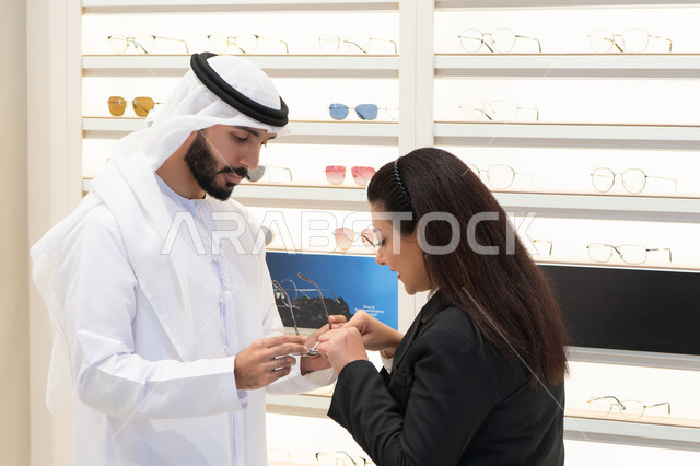 Admiring eyeglasses while shopping, buying accessories from malls, an Emirati woman helping customers choose the right sizes for them, taking care of their appearance, the concept of weak eyesight, an Emirati man wearing a kandura and a white ghutra choosing the right glasses
