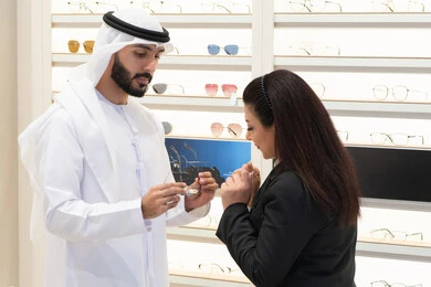 Buying new eyeglasses, buying accessories in malls, an Emirati woman helping customers choose the right sizes for them, taking care of their appearance, the concept of weak eyesight, an Emirati man wearing a white kandura and ghutra choosing the right glasses