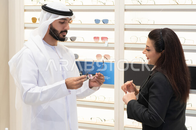 Buying accessories in malls, admiring glasses while shopping, an Emirati woman helping customers choose the right sizes for them, taking care of their appearance, the concept of weak eyesight, an Emirati man wearing a kandura and a white ghutra choosing the right glasses