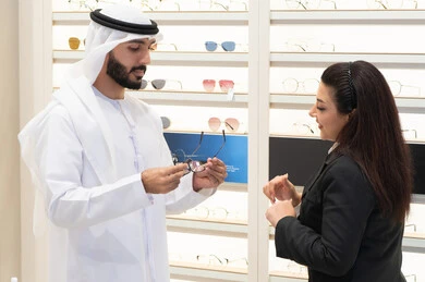 Buying accessories in malls, admiring glasses while shopping, an Emirati woman helping customers choose the right sizes for them, taking care of their appearance, the concept of weak eyesight, an Emirati man wearing a kandura and a white ghutra choosing the right glasses