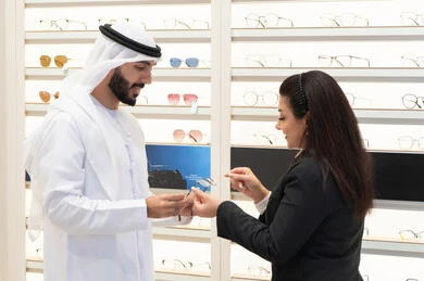 Admiring glasses while shopping, an Emirati woman helping customers choose the right sizes for them, shopping in an Emirati mall, taking care of one's appearance, the concept of weak eyesight, an Emirati man wearing a kandura and a white ghutra choosing the right glasses