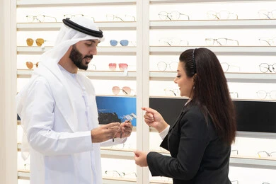 Glasses display shelves, Emirati woman helping customers choose the right size, shopping in an Emirati mall, taking care of one's appearance, the concept of weak eyesight, Emirati man wearing a white kandura and ghutra choosing the right glasses