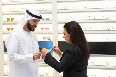 Emirati woman helping customers choose the right sizes for them, shopping in an Emirati mall, taking care of their appearance, buying eyeglasses, the concept of weak eyesight, Emirati man wearing a kandura and white ghutra choosing the right glasses