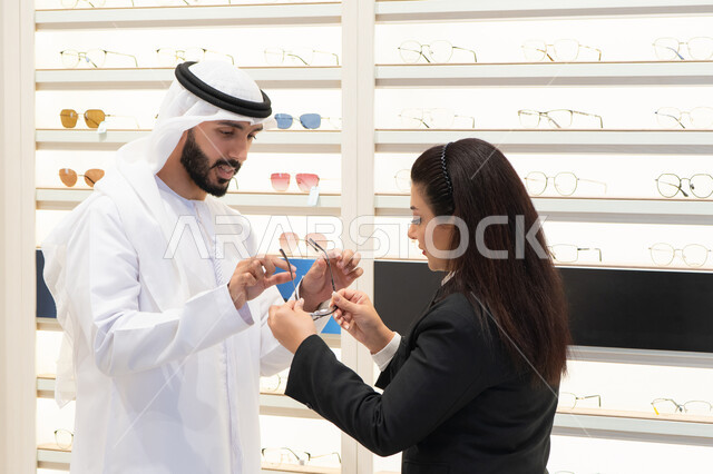 التسوق باحدى المولات الاماراتيه ، الاهتمام بالمظهر الخارجي، شراء النظارات الطبية، مفهوم ضعف النظر، رجل اماراتي يرتدي الكندوره والغتره البيضاء يقوم باختيار النظارة المناسبة، سيده اماراتيه تقوم بمساعده الزبائن في اختيار المقاسات المناسبه لهم