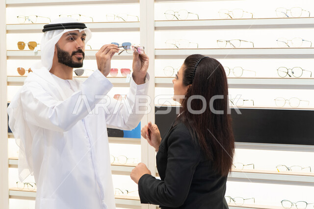 Caring about one's appearance, buying eyeglasses, the concept of weak eyesight, shopping in malls, an Emirati man wearing a white kandura and ghutra choosing the right glasses, an Emirati woman helping customers choose the right sizes for them
