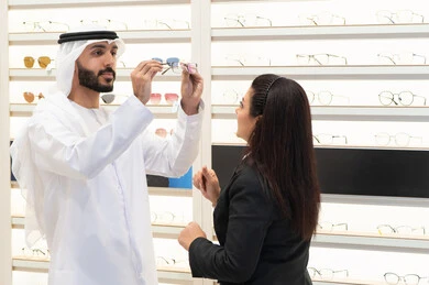 Caring about one's appearance, buying eyeglasses, the concept of weak eyesight, shopping in malls, an Emirati man wearing a white kandura and ghutra choosing the right glasses, an Emirati woman helping customers choose the right sizes for them