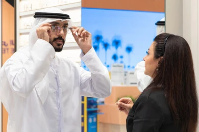 Emirati man wearing white kandura and ghutra choosing suitable glasses, glasses display shelves in a store, shopping in malls, concept of weak eyesight, Emirati woman helping customers choose the appropriate sizes for them, taking care of external appearance