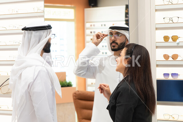 Glasses display shelves in a store, shopping in malls, the concept of poor eyesight, an Emirati woman helping customers choose the appropriate sizes for them, taking care of their appearance, an Emirati man wearing a white kandura and ghutra choosing the appropriate glasses