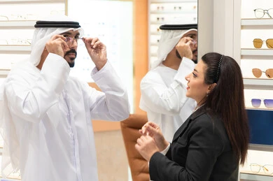 Shopping and buying from malls, the concept of weak eyesight, glasses display shelves in a store, an Emirati woman helping customers choose the appropriate sizes for them, taking care of their appearance, an Emirati man wearing a kandura and a white ghutra choosing the appropriate glasses