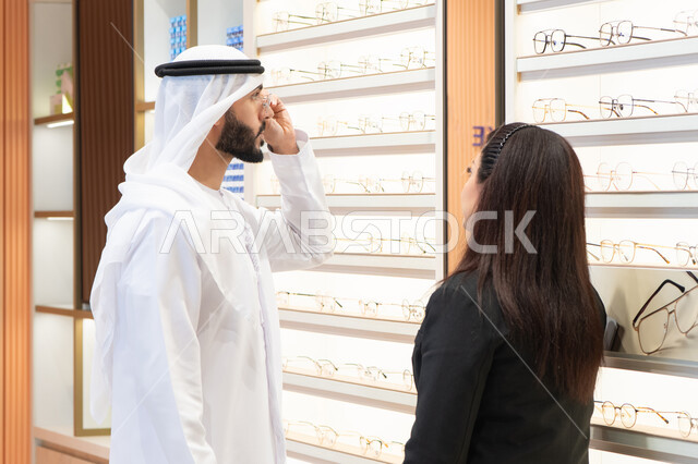 Shopping for prescription glasses from a mall. An Emirati woman helps customers choose the right size. Taking care of one's appearance. An Emirati man wearing a kandora and a white ghutra selects the right pair of prescription glasses. The concept of poor vision.