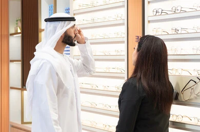 Shopping for prescription glasses from a mall. An Emirati woman helps customers choose the right size. Taking care of one's appearance. An Emirati man wearing a kandora and a white ghutra selects the right pair of prescription glasses. The concept of poor vision.