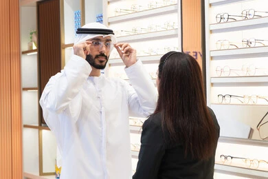 Emirati woman helping customers choose the right sizes, shopping for eyeglasses from malls, taking care of their appearance, Emirati man wearing white kandura and ghutra choosing the right eyeglasses, following the latest fashion trends, concept of weak eyesight