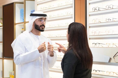 Shopping and buying eyeglasses from malls, taking care of one's appearance, an Emirati man wearing a white kandura and ghutra choosing the right eyeglasses, following the latest fashion trends, an Emirati woman helping customers choose the right sizes for them, the concept of weak eyesight