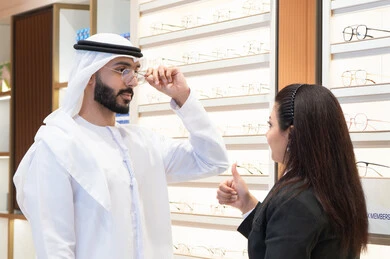 Emirati man wearing white kandura and ghutra choosing suitable eyeglasses, following the latest fashion trends, Emirati woman helping customers choose the right sizes for them, concept of weak eyesight, taking care of appearance, shopping and buying eyeglasses from malls
