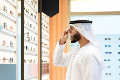 Emirati man wearing white kandura and ghutra buying suitable sunglasses in an optical store, following the latest fashion and beauty trends, shopping and buying the latest modern eyewear products, the corner for luxury sunglasses, taking care of one's appearance