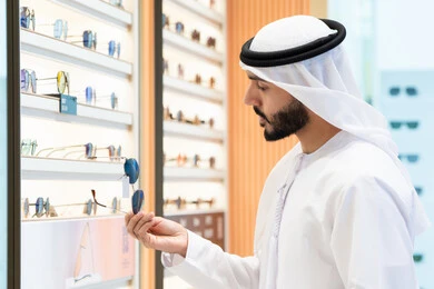 متابعة آخر صيحات الموضة والجمال, تسوق وشراء أحدث المنتجات العصرية الحديثة, الركن الخاص بالنظارات الشمسيه الفخمه ،الاهتمام بالمظهر الخارجي، رجل اماراتي يرتدي الكندوره والغتره البيضاء يقوم بشراء النظارة المناسبة في محل اماراتي