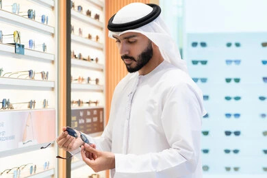 التسوق والتبضع من المولات، الاهتمام بالمظهر الخارجي، الركن الخاص بالنظارات الشمسيه الفخمه ،رجل اماراتي يرتدي الكندوره والغتره البيضاء يقوم بشراء النظارة المناسبة في محل اماراتي