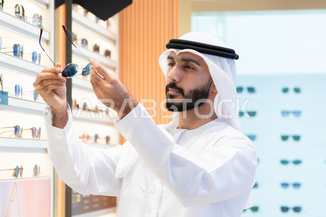 الركن الخاص بالنظارات الشمسيه الفخمه ،الاهتمام بالمظهر الخارجي، رجل اماراتي يرتدي الكندوره والغتره البيضاء يقوم بشراء النظارة المناسبة في محل اماراتي ،  التسوق والتبضع من المولات