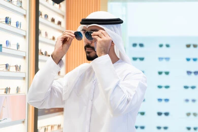 رجل عربي خليجي اماراتي يرتدي الكندوره يقوم بقياس نظاره سوداء، ارفف عرض النظارات داخل المحل، ايماءات الثقه في النفس ،الفحص قبل الشراء، متعة التسويق والتبضع باختيار نظارة شمسيه مناسبة