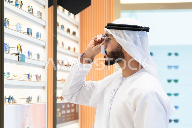 ارفف عرض النظارات داخل المحل، ايماءات الثقه في النفس ،الفحص قبل الشراء، متعة التسويق والتبضع باختيار نظارة شمسيه مناسبة، رجل عربي خليجي اماراتي يرتدي الكندوره يقوم بقياس نظاره سوداء