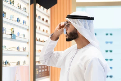 Eyeglasses display shelves inside a store, gestures of self-confidence, inspection before purchase, the joy of shopping and choosing suitable sunglasses, an Emirati Gulf Arab man wearing a kandura trying on a pair of black sunglasses.