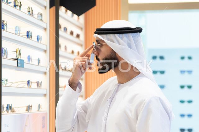 Types and brands of international glasses, gestures of self-confidence, examination before purchase, the joy of shopping and choosing suitable sunglasses, an Emirati Gulf Arab man wearing a kandura trying on a pair of black sunglasses.