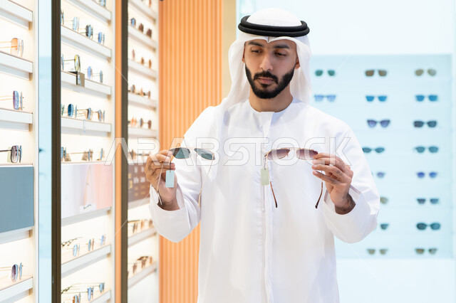 Looking at a collection of frames inside a mall, sunglasses store, buying sun protection glasses, an Arab Gulf Emirati man wearing a kandura and ghutra choosing the right one in an optical store