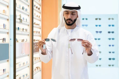 Looking at a collection of frames inside a mall, sunglasses store, buying sun protection glasses, an Arab Gulf Emirati man wearing a kandura and ghutra choosing the right one in an optical store