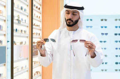 An Emirati Gulf Arab man wearing a kandura and ghutra choosing the right pair of sunglasses in an optical shop, looking at a collection of frames inside a mall, a sunglasses exhibition, buying sun protection glasses