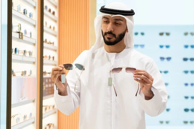 Sunglasses exhibition, the pleasure of shopping, an Emirati Gulf Arab man wearing a kandura and ghutra choosing the appropriate one in an Emirati store, confusion and choosing between two glasses, looking at a group of frames inside the mall