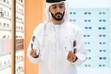 اقتناء نظارات حماية من الشمس، رجل عربي خليجي اماراتي يرتدي الكندورة والغترة يقوم باختيار المناسب في محل اماراتي، الحيره والاختيار بين نظارتين ،النظرعلى مجموعة من الاطارات داخل المول ، تشكيلة نظارات شمسيه أنيقة