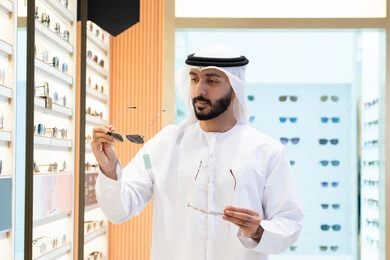 An Emirati Gulf Arab man wearing a kandura and ghutra choosing the right pair of sunglasses in an Emirati store, confused and choosing between two pairs of sunglasses, looking at a selection of frames inside a mall, wearing and choosing sunglasses, a selection of stylish glasses.