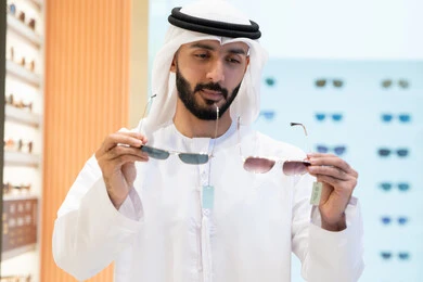 Confusion and choosing between two pairs of glasses, looking at a collection of frames inside a mall, wearing and choosing sunglasses, a selection of elegant glasses, an Emirati Gulf Arab man wearing a kandura and ghutra choosing the right pair in an Emirati store.