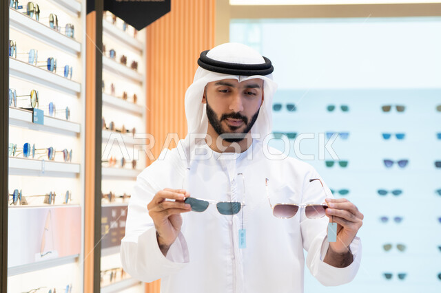 النظر على مجموعة من الاطارات داخل المول ، ارتداء و اختيار النظاره ، تشكيلة نظارات شمسية أنيقة ،رجل عربي خليجي اماراتي يرتدي الكندورة والغترة يقوم باختيار المناسب في محل اماراتي