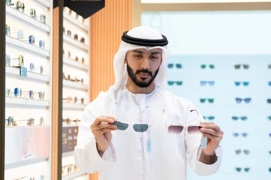 Sunglasses exhibition, shopping fun, an Emirati Gulf Arab man wearing a kandura and ghutra choosing the right pair in an Emirati store, confusion and choosing between two pairs of glasses, looking at a collection of frames inside a mall.  Looking at a collection of frames inside a mall, wearing and choosing glasses, a selection of stylish sunglasses, an Emirati Gulf Arab man wearing a kandura and ghutra choosing the right pair in an Emirati store.