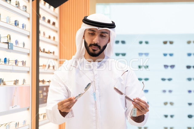 تشكيلة نظارات شمسية أنيقة ، ارتداء و اختيار النظاره الشمسيه، رجل عربي خليجي اماراتي يرتدي الكندورة والغترة يقوم باختيار المناسب في محل اماراتي, النظرعلى مجموعة من الاطارات داخل المول